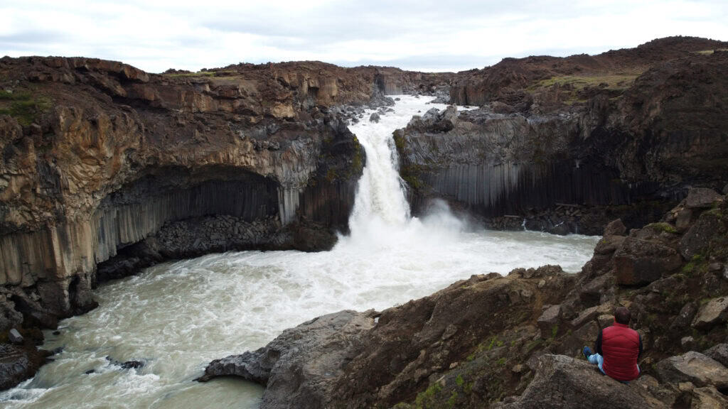 Photographer sitting on basalt rock overlooking Aldeyjarfoss waterfall in Iceland's northern highlands — hexagonal basalt columns and glacial river with no other visitors in sight