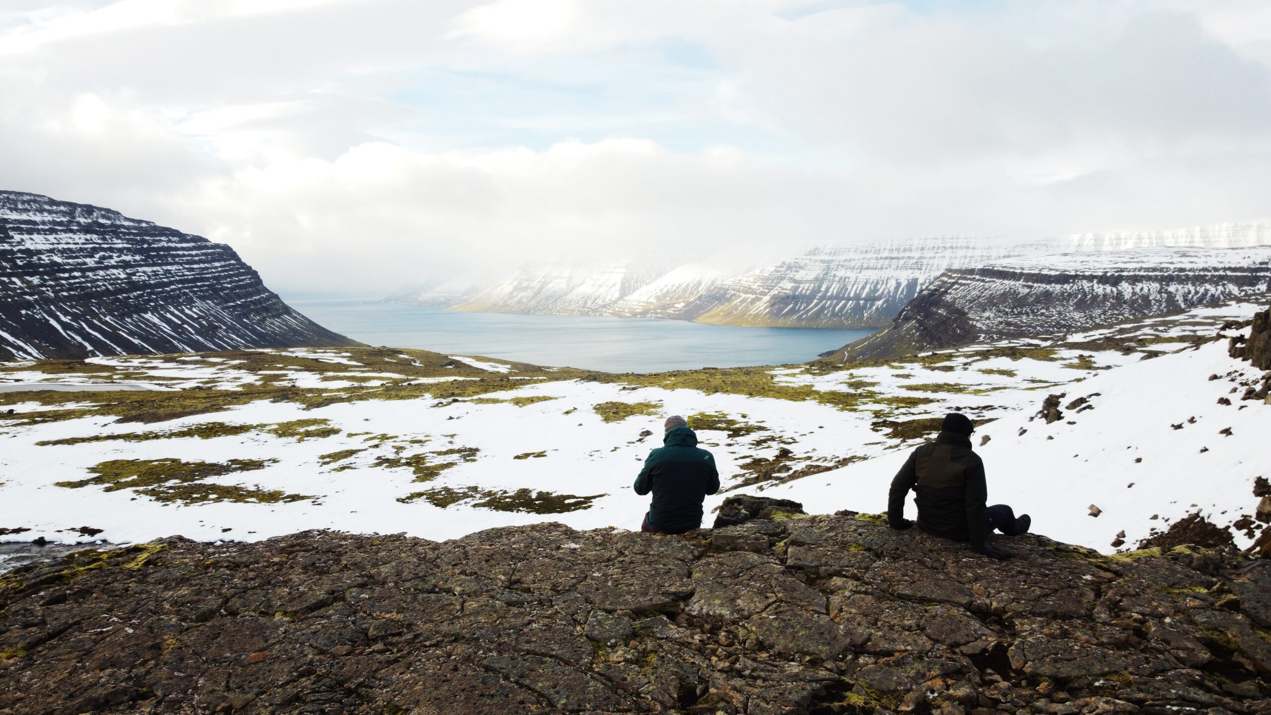 Aerial view over a Westfjords fjord, Iceland, with two photographers resting on the rocks — photographed by Marcel Strobel