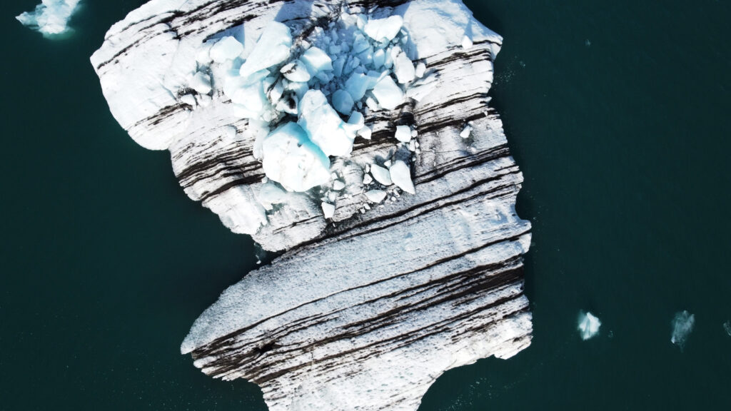 Aerial drone view of a striped glacial iceberg floating in glacier lagoon, Iceland — showing the layered sediment bands only visible from above