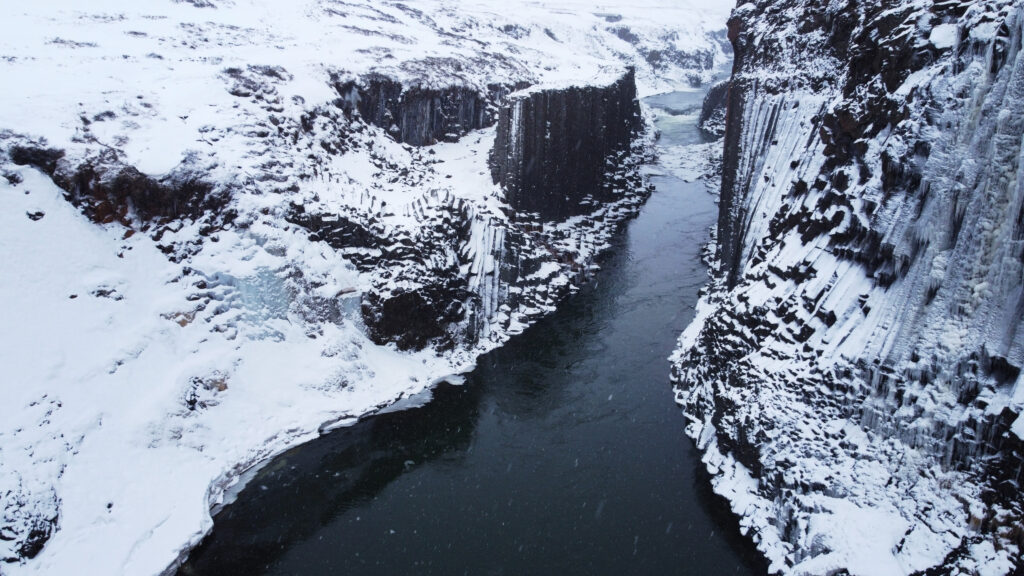Aerial view of a snow-covered basalt column canyon in Iceland in winter, river flowing through frozen gorge walls — drone photography revealing inaccessible terrain

