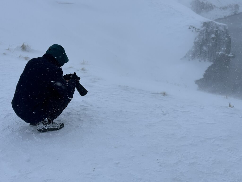 Photographer crouching in a blizzard in Iceland, shooting hand-held in heavy snow — demonstrating why weather-sealed equipment matters in Iceland's conditions

