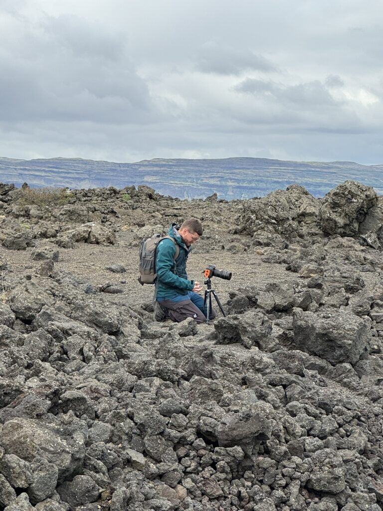 Photographer with camera backpack and tripod working on a rough lava field in Iceland — showing the physical demands of carrying equipment across Iceland's terrain


