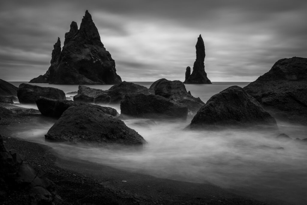 Black and white long exposure of Reynisdrangar sea stacks at Reynisfjara beach, south Iceland — volcanic rock formations in silky surf under a dramatic overcast sky
