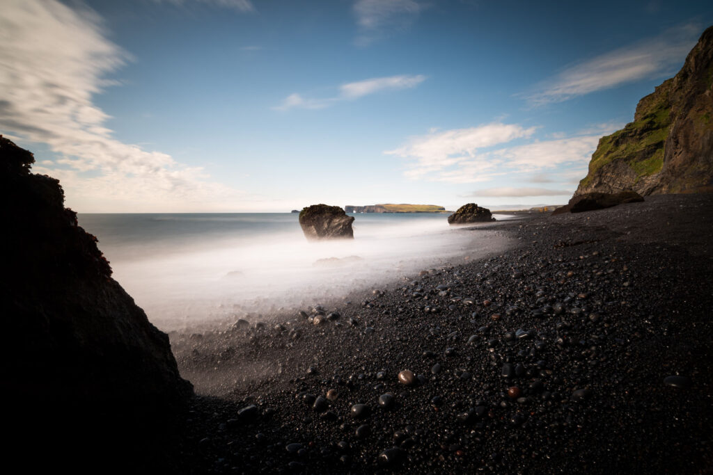Long exposure photograph of black sand beach at Reynisfjara, Iceland — silky wave motion between volcanic rocks with Reynisdrangar sea stacks and Dyrhólaey peninsula visible in the background

