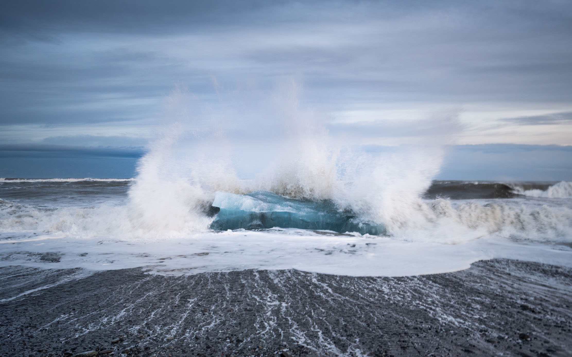 Glacial ice block at Diamond Beach, Iceland, engulfed by an Atlantic wave on black volcanic sand — photographed by Marcel Strobel