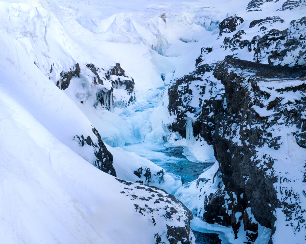 Frozen river canyon in Iceland in winter, with turquoise glacial water flowing beneath ice formations and deep snow — a winter landscape only accessible in the cold season
