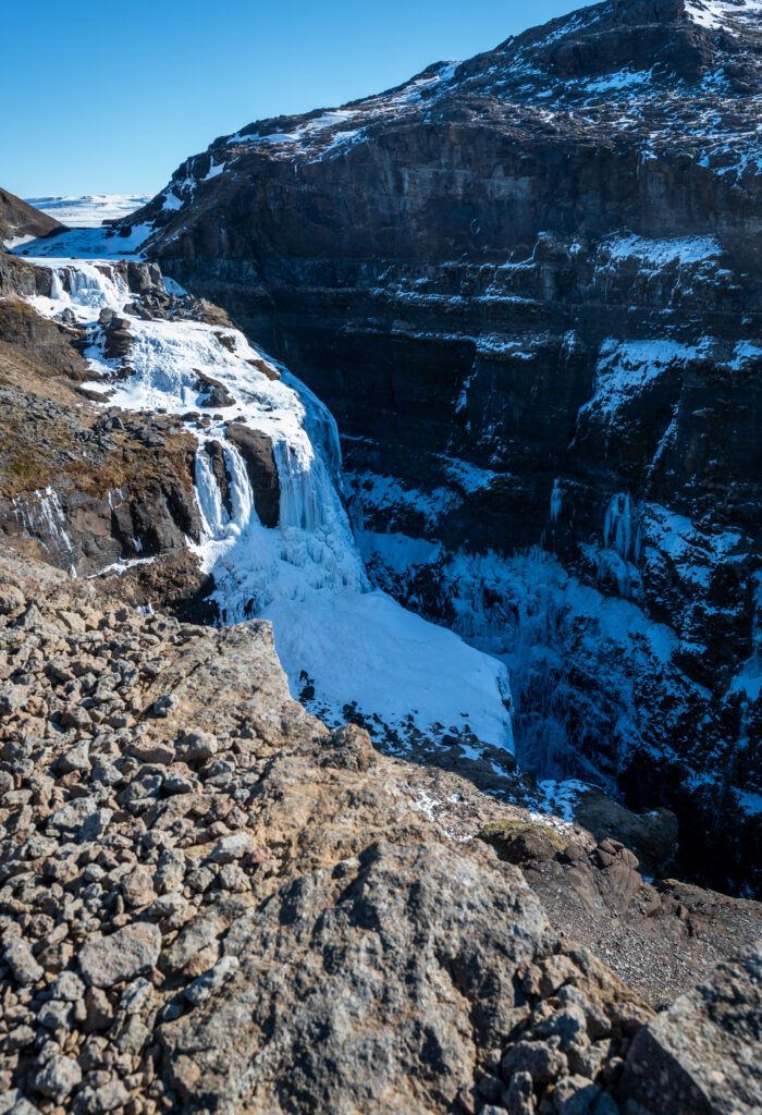 Partially frozen waterfall in Iceland in winter, with a lone figure visible on the ice field below — showing the dramatic scale of Iceland's frozen waterfalls accessible only in the cold season