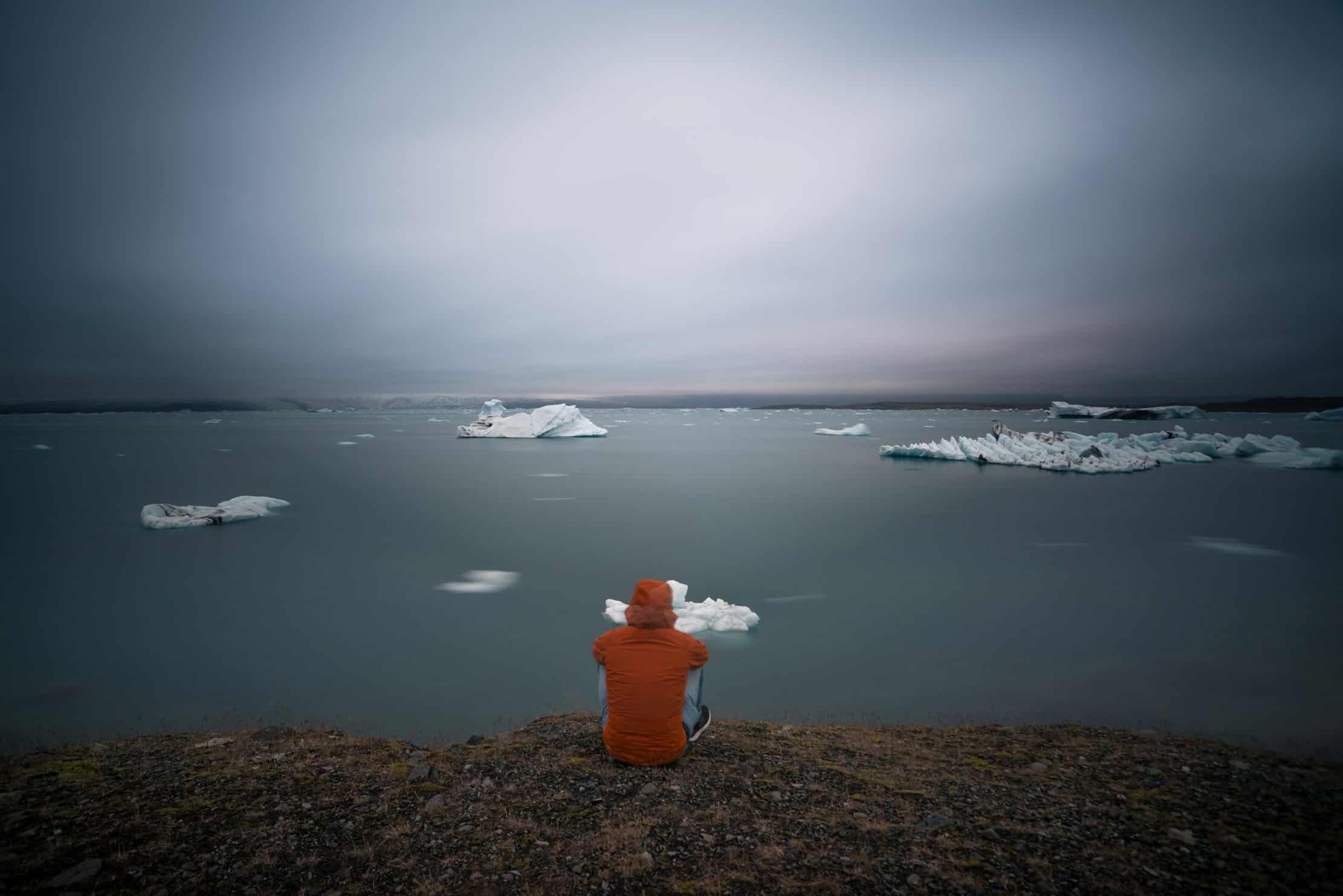 Photographer sitting at the shore of Jökulsárlón glacier lagoon, Iceland, watching icebergs drift in still water under a dramatic overcast sky