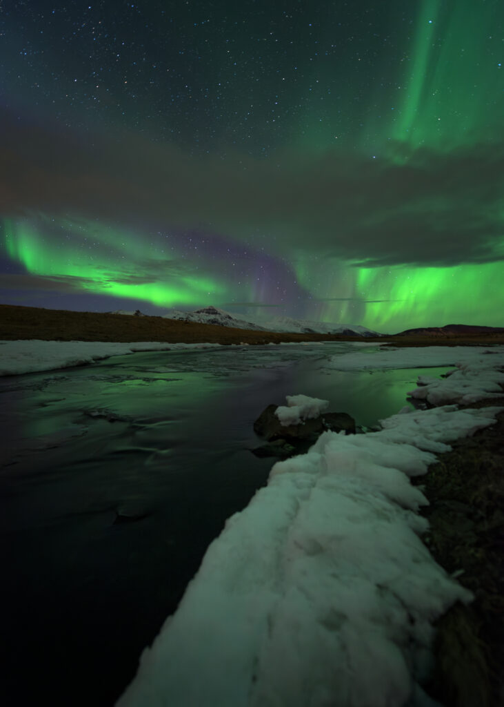 Northern lights with green and purple aurora reflected in a snow-edged river in Iceland, with snow-capped mountains on the horizon — KP 4–5 display