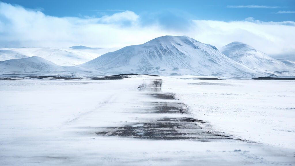 Snow-covered road through a white winter landscape in Iceland, barely visible under drifting snow with mountains in the background — illustrating the serious driving conditions photographers face in winter