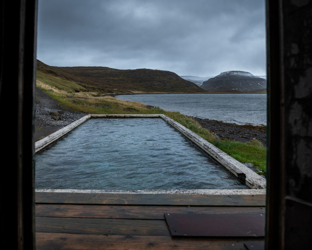 Geothermal hot pool in the Westfjords, Iceland, framed by a wooden doorway with a view across a remote fjord and snow-capped mountains

