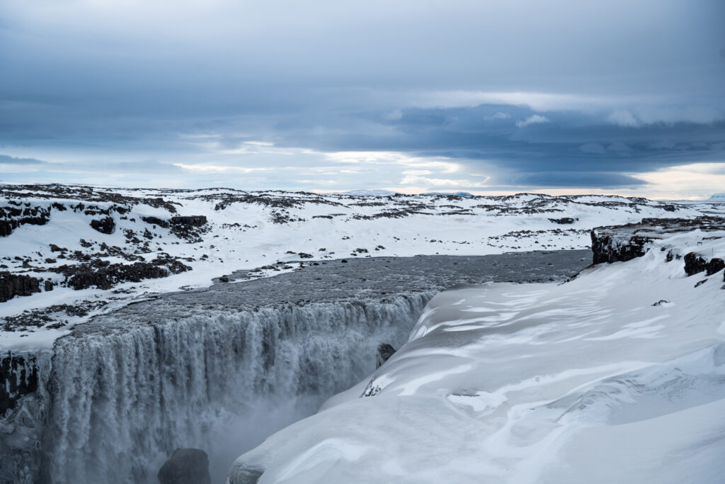 Dettifoss waterfall in winter from the west bank, Iceland — partially frozen falls against a snow-covered lava plateau, accessible year-round via the paved Route 862
