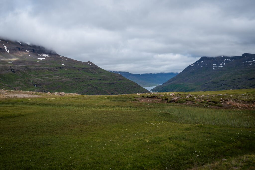 View from the Route 93 mountain pass descent toward Seyðisfjörður, East Iceland — green valley with the fjord visible at the far end, waterfalls cascading from the hillsides on both sides