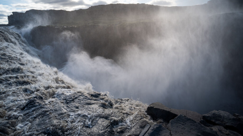 Dettifoss waterfall from the east bank rim, Iceland — glacial water rushing over the edge with spray filling the Jökulsárgljúfur canyon, tiny figures visible on the west bank platform in the background