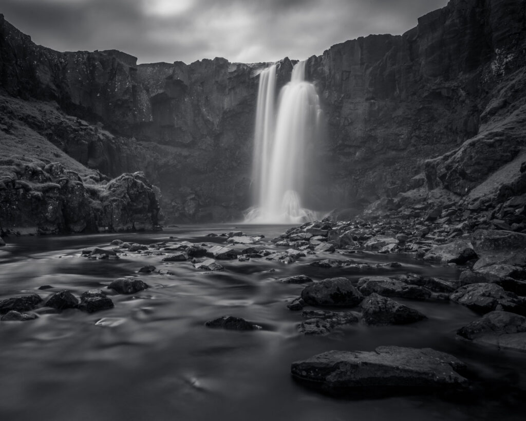 Long exposure black and white photograph of a waterfall above Seyðisfjörður, East Iceland — smooth water column between basalt canyon walls with rocky foreground
