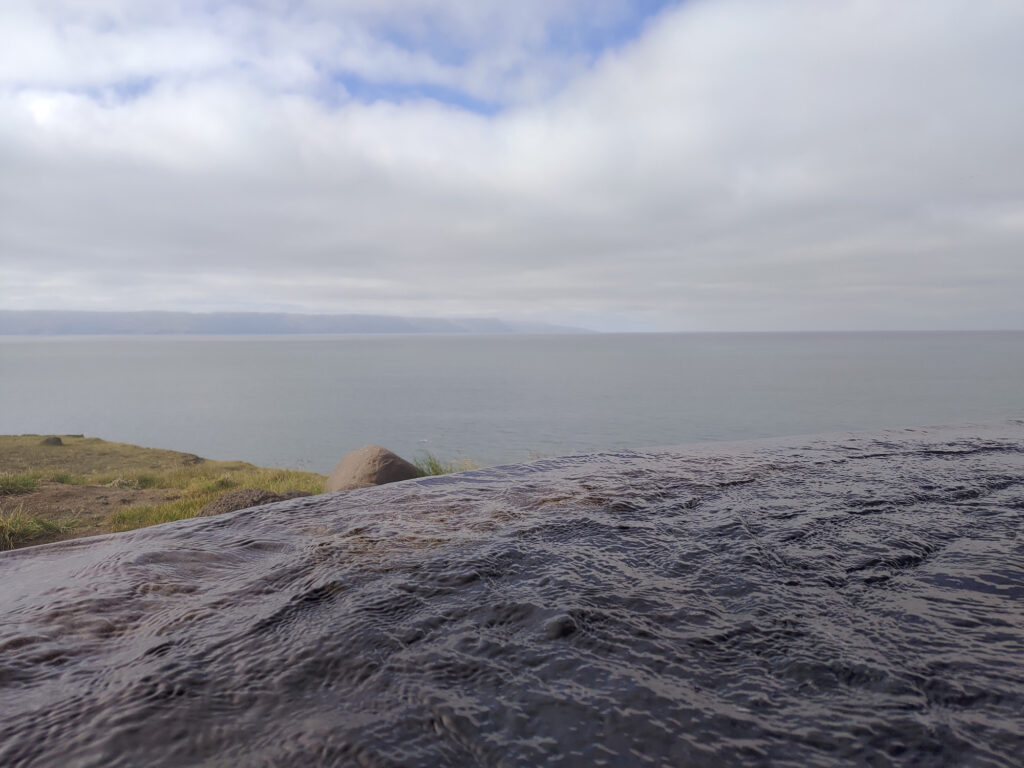 Close-up of warm geothermal water flowing over dark volcanic rock with the ocean and distant coastline visible behind, Iceland — texture and context rather than a specific pool