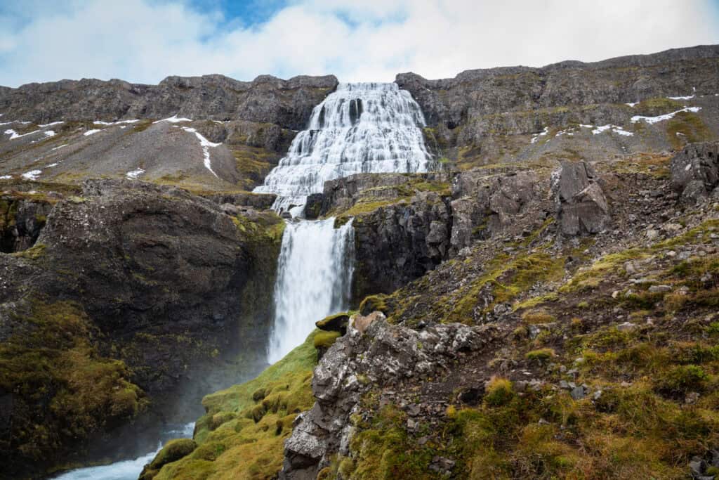 Dynjandi waterfall in the Westfjords, Iceland, in autumn — the tiered cascade widening toward the base, surrounding hillsides with first snow, photographed in October