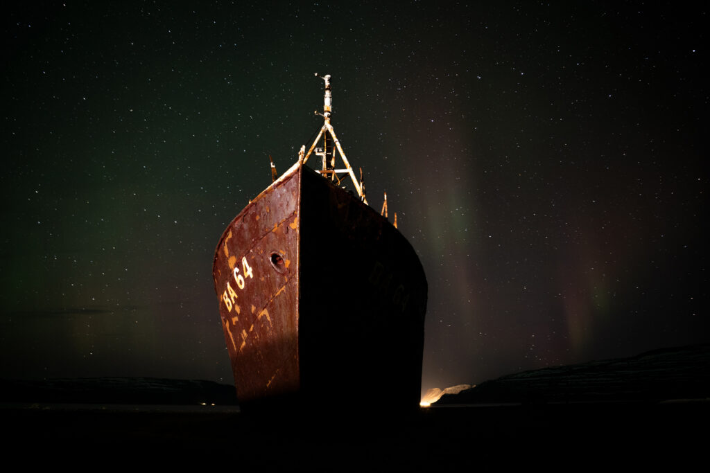 Garðar BA 64 shipwreck bow at night under the Northern Lights, Patreksfjörður, Westfjords, Iceland — rusted hull lit from below with aurora columns visible on both sides and a clear star-filled sky above