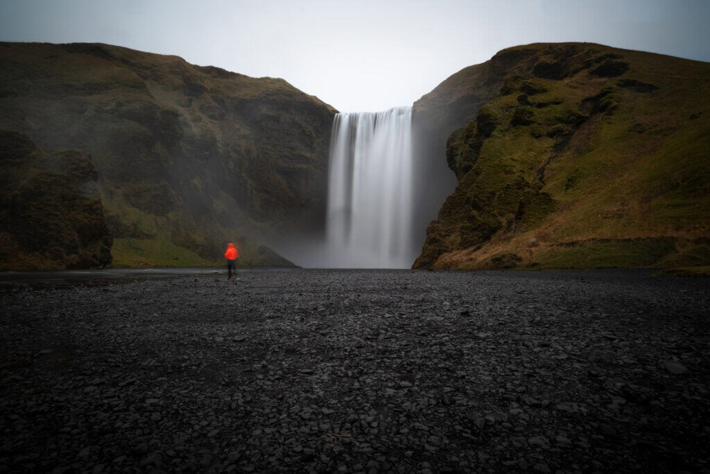 Long exposure of Skógafoss waterfall in Iceland with a lone figure in a red jacket in the foreground — the human scale revealing the true height of the falls, black volcanic gravel in the foreground, overcast winter light