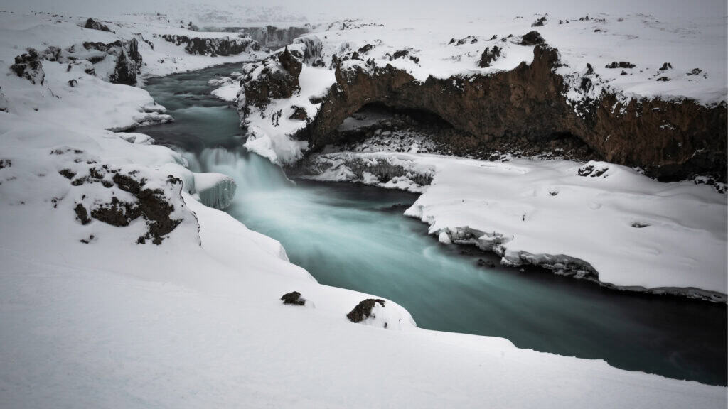 Long exposure of the river above Goðafoss waterfall in winter, Iceland — turquoise glacial water flowing between snow-covered lava formations with partial ice formations along the banks