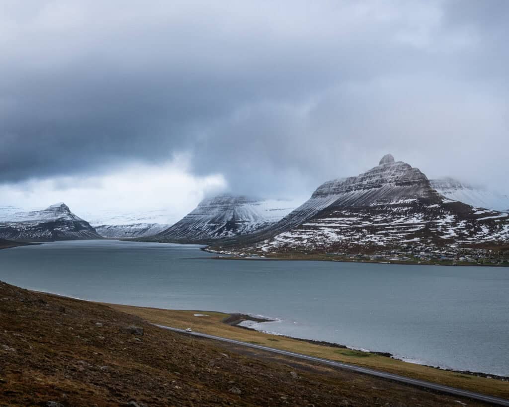 A single car on a coastal road in the Westfjords of Iceland, snow-covered table mountains rising on both sides of the fjord under a stormy overcast sky