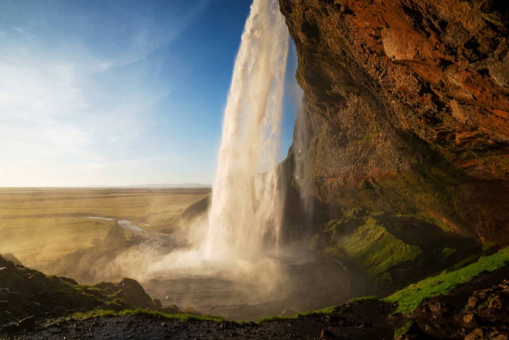 Seljalandsfoss waterfall in Iceland, photographed from the cave behind the falls at golden hour, with the South Coast plains visible in the background